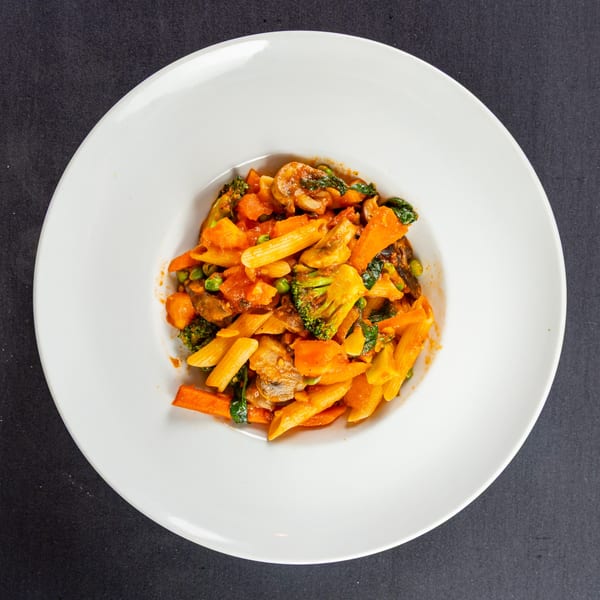 Penne pasta with broccoli, carrots, mushrooms, and peas in a rich tomato sauce on a white plate.