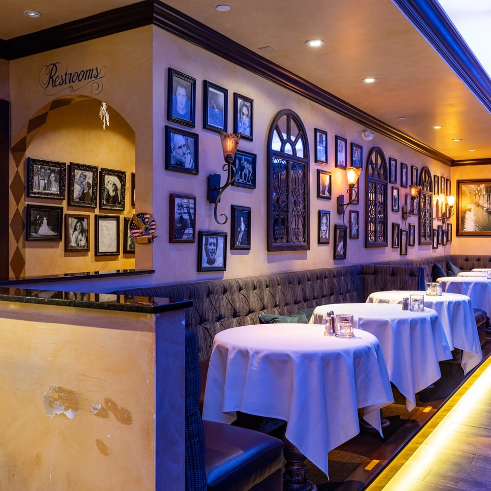 Cozy Italian restaurant interior with framed photos, sconces, and white-clothed tables along a banquette.